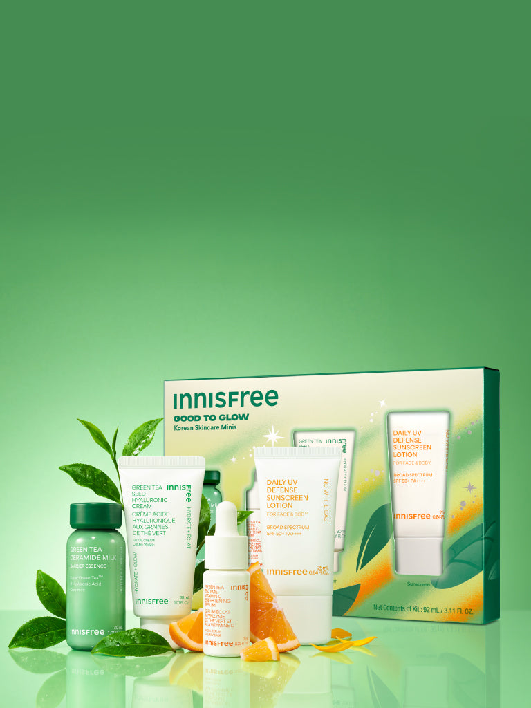 Innisfree Good to Glow skincare set displayed with its retail box on a reflective light green surface. The set includes a Green Tea Ceramide Milk mini, Green Tea Hyaluronic Cream mini, Vitamin C Serum mini, and Daily UV Sunscreen SPF 50+ mini, styled with green leaves and orange slices.