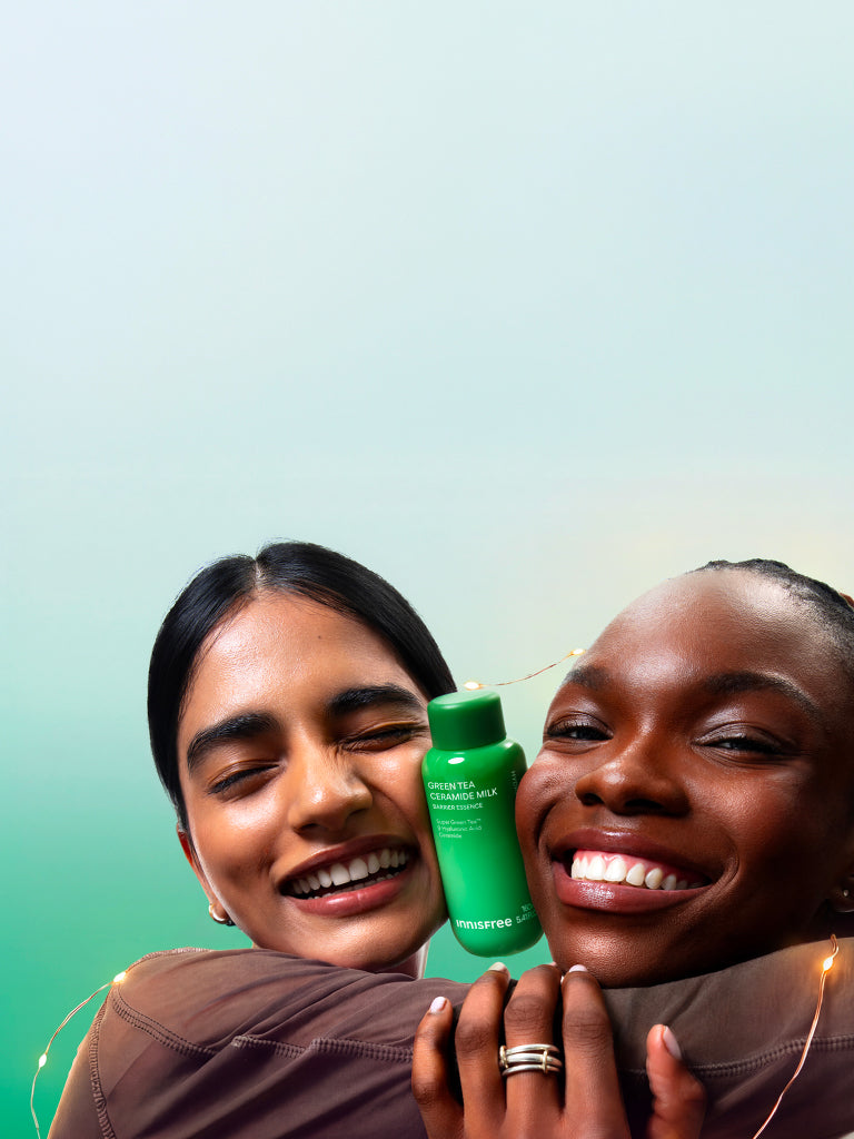 Two smiling women hugging while holding a green bottle of Green Tea Ceramide Milk. The background is a soft gradient of light blue to green with warm string lights visible around them.