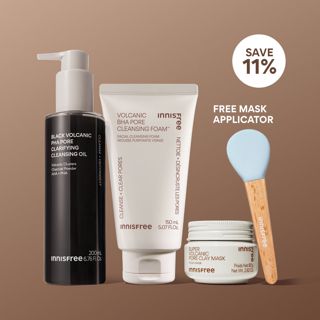 The Volcanic Pore Care Routine featuring Black Volcanic PHA Pore Cleansing Oil, Volcanic BHA Pore Cleansing Foam, two light blue silicone cleansing brushes with wooden handles, and a Super Volcanic Pore Clay Mask jar, arranged on a solid brown background: save 11% and free mask applicator