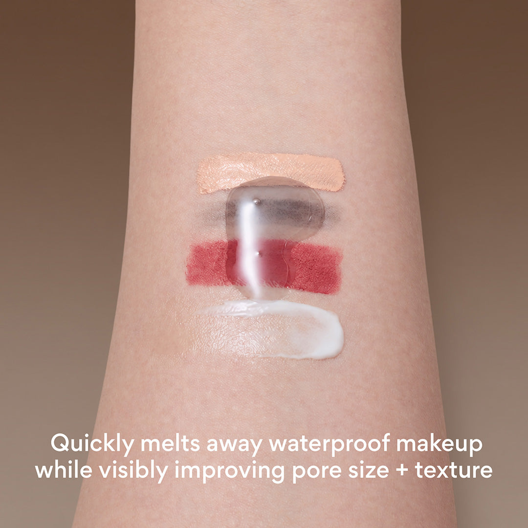Close-up of a person's forearm with four makeup swatches applied: a beige cream, mascara smudge, a red lipstick layer, and a white cream. A translucent clear product is being pressed over the center, with text at the bottom reading 'Quickly melts away waterproof makeup while visibly improving pore size + texture'.