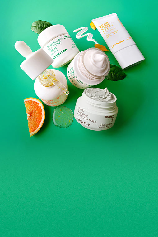 A flat-lay arrangement of Immsree skincare products on a bright green background, including white jars, a dropper bottle, a tube, and a peel, styled with green leaves, an orange slice, and a green gel cube.
