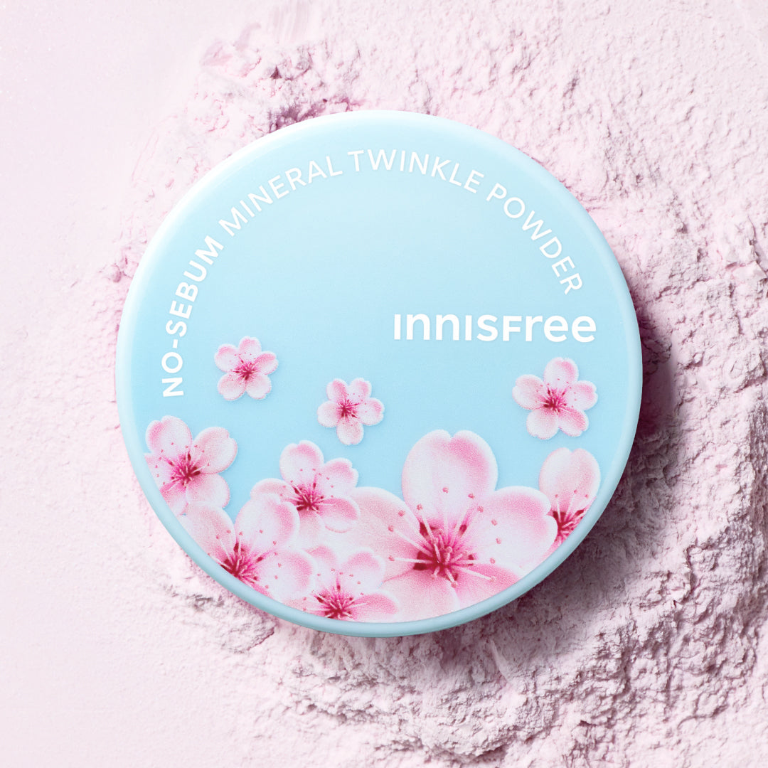 A round compact of Innisfree No-Sebum Mineral Twinkle Powder Cherry Blossom, featuring a light blue lid decorated with pink cherry blossoms, set against a background of loose pink powder.