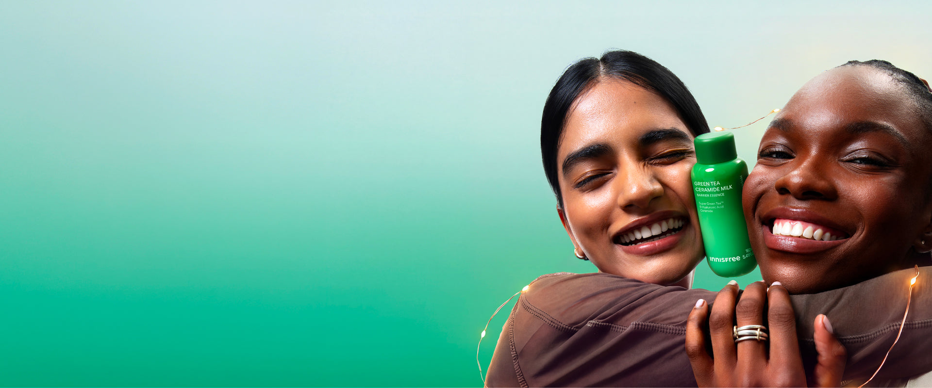 Two smiling women hugging while holding Green Tea Ceramide Milk against a soft teal gradient background.