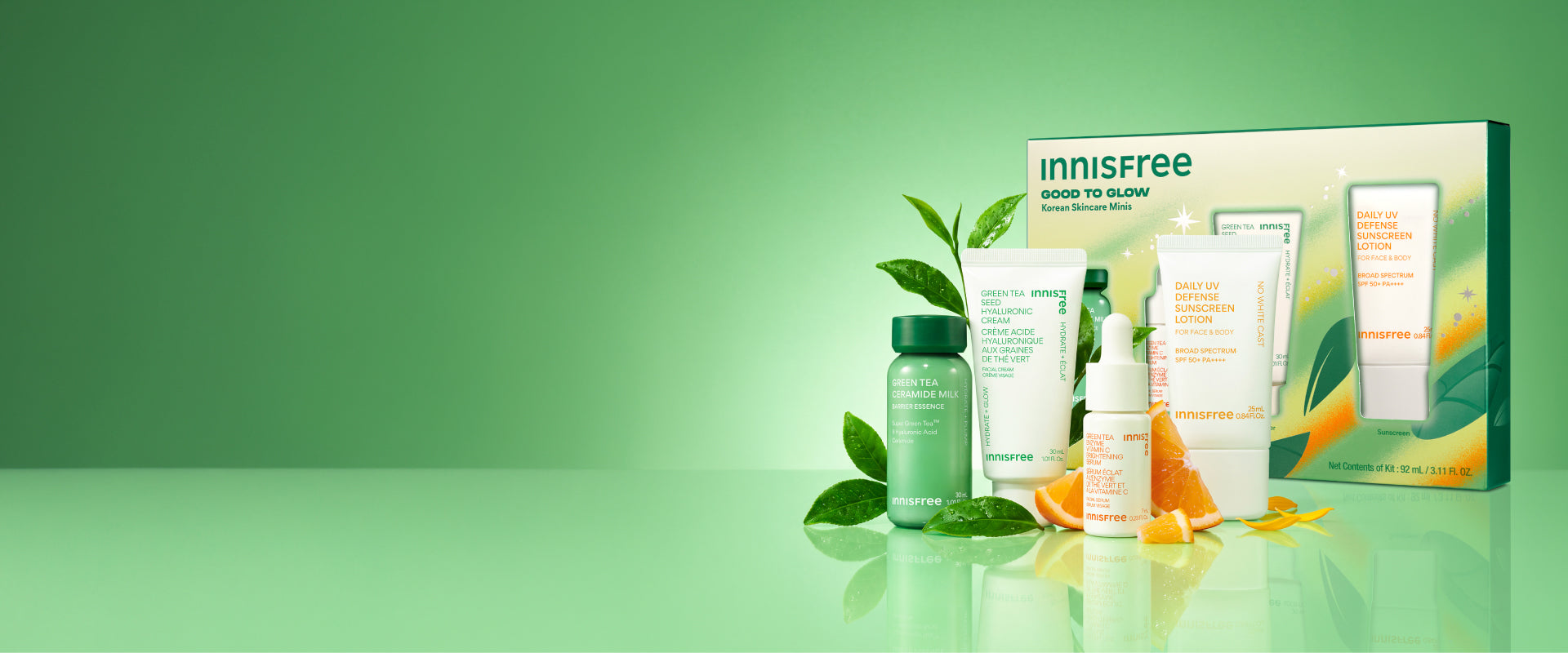 Innisfree Good to Glow skincare set displayed with its retail box on a reflective light green surface. The set includes a Green Tea Ceramide Milk mini, Green Tea Hyaluronic Cream mini, Vitamin C Serum mini, and Daily UV Sunscreen SPF 50+ mini, styled with green leaves and orange slices.