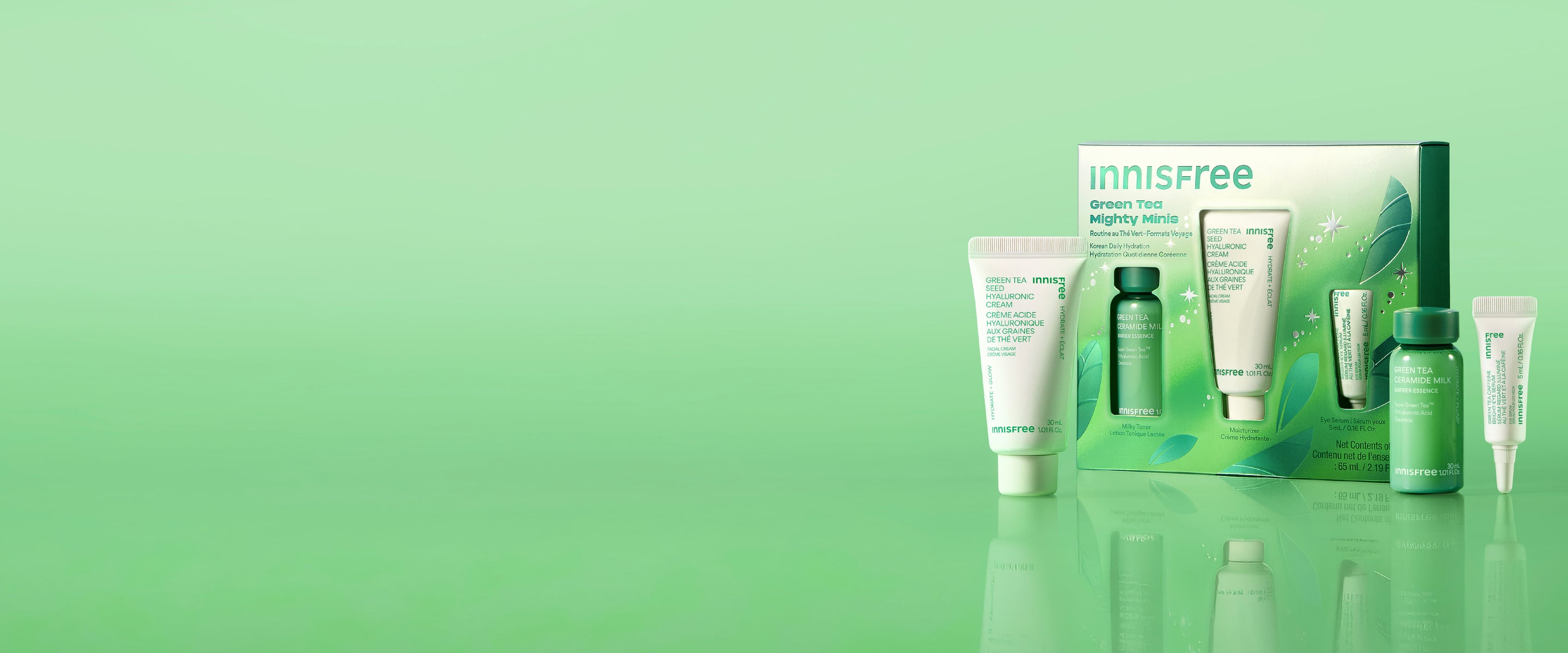 Green Tea Mighty Minis set with skincare tubes and bottles in white and green packaging, displayed on a reflective green surface against a light green gradient background.