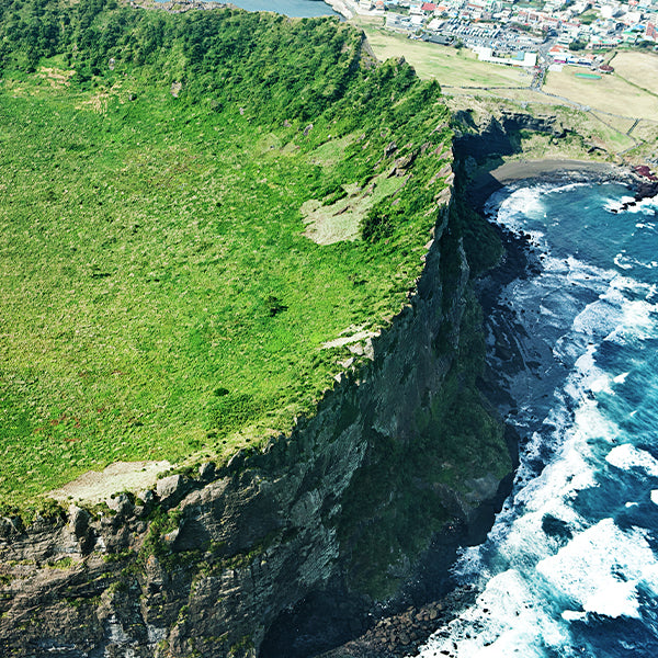 Aerial view of a steep green cliff overlooking a rocky ocean shoreline with white surf, a small coastal town at the top, and a dark shadowed cave opening along the cliff edge in Jeju