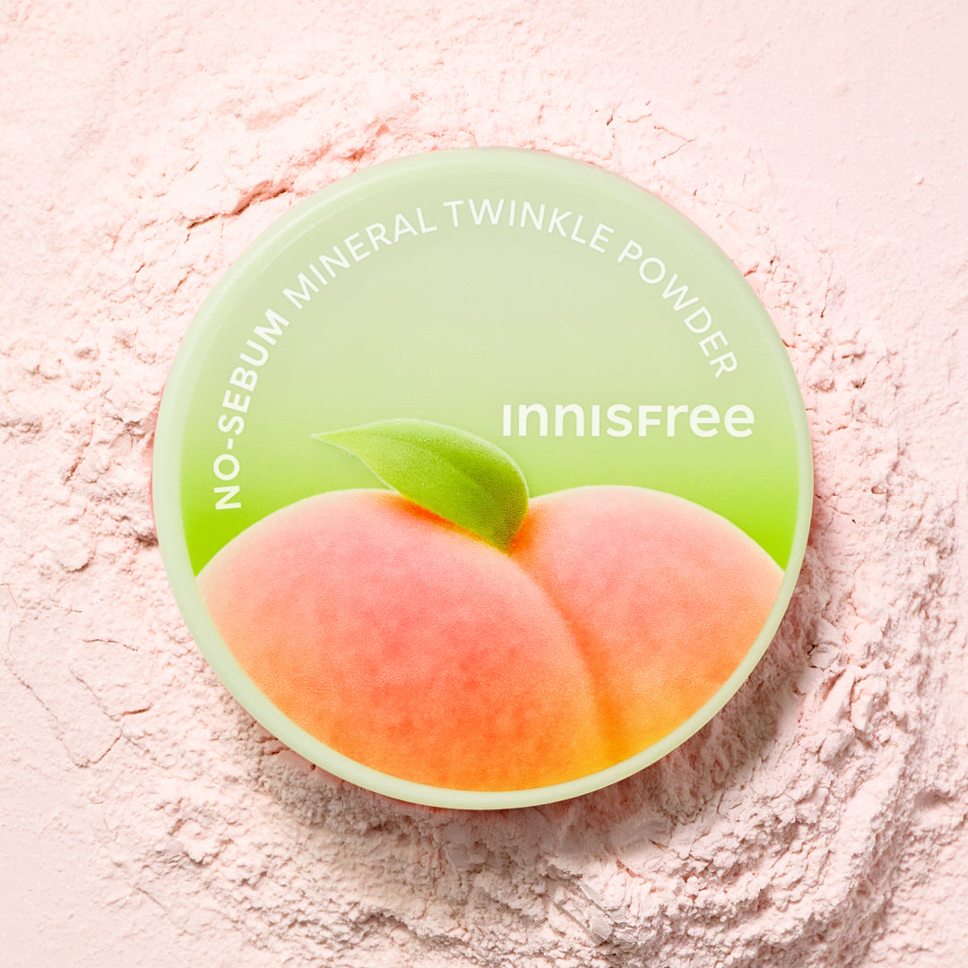 Innisfree No-Sebum Mineral Twinkle Powder Peach Blossom compact lid featuring a peach illustration on a light green background, set against a bed of pink cosmetic powder.