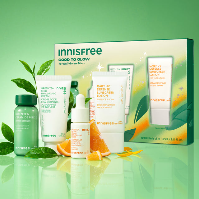 Innisfree Good to Glow skincare set displayed with its retail box on a reflective light green surface. The set includes a Green Tea Ceramide Milk mini, Green Tea Hyaluronic Cream mini, Vitamin C Serum mini, and Daily UV Sunscreen SPF 50+ mini, styled with green leaves and orange slices.